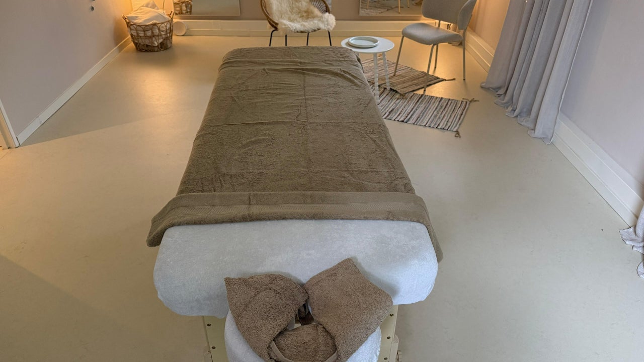 Zicht over de massagetafel naar de rusthoek met stoelen en een staande lamp